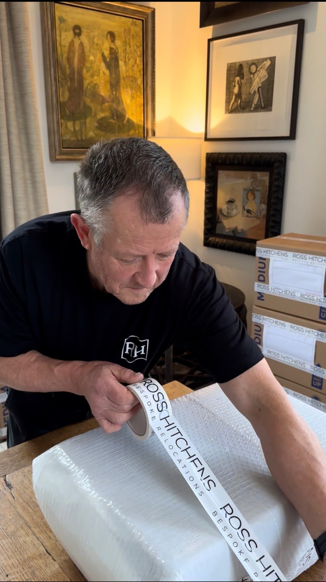Ross Hitchens wrapping an item in front of framed artwork, with packed cartons beside him